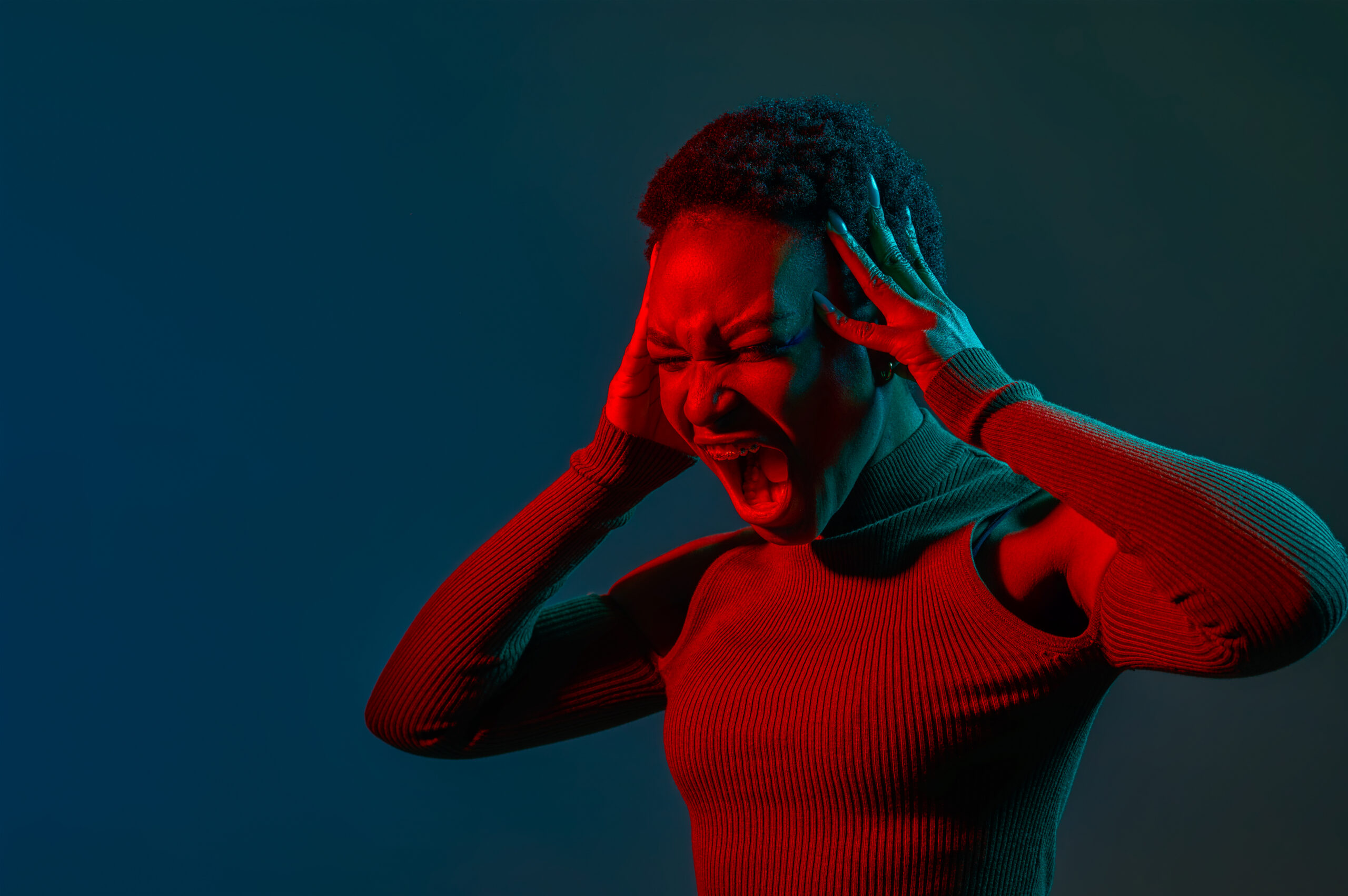 Headshot portrait of emotional young woman crazy shouting holding head with hand in neon light over dark studio background