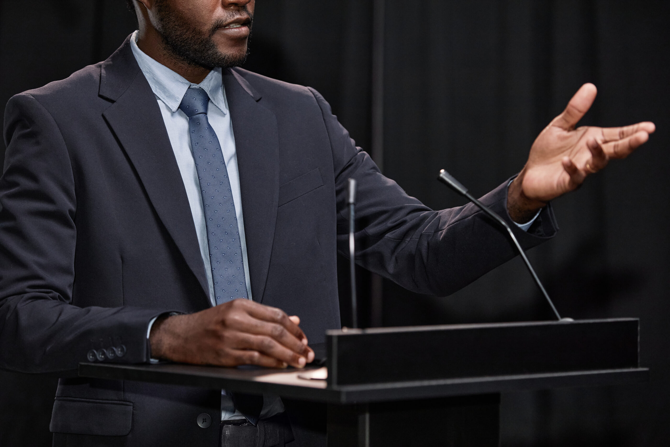 Cropped shot of African American male politician dressed in blue suit gesticulating with hands speaking to audience, while giving public speech on stage at podium against black curtains